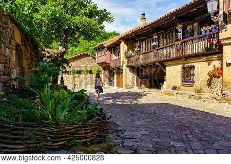 Woman Walking Through Village Of Old Houses With Balconies With Flowers. Barcena Mayor Santander.