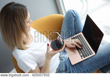 Young Woman Sitting On Armchair With Laptop And Drinking Red Wine From Glass