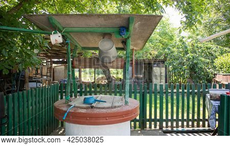 An Old Water Well With A Bucket In The Garden, A Metal Handle For Lifting Water From The Ground. Vil