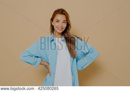 Self Assured Smiling Lovely Model Keeps Hands On Waist, Wears Long Blue Shirt Over White Tshirt, Loo