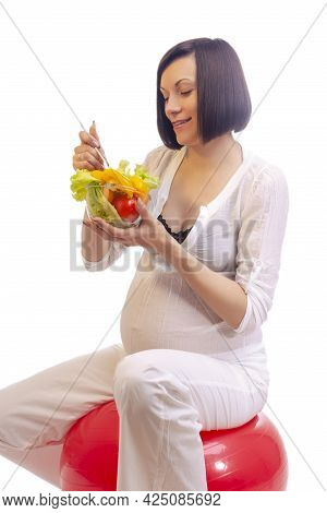 Smiling Pregnant Brunette Woman Sitting On Fitball And Eating Fresh Vegetables In Glass Bowl Over Wh