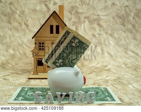 A Piggy Bank With A Dollar Stands On A Dollar Near A Wooden House.