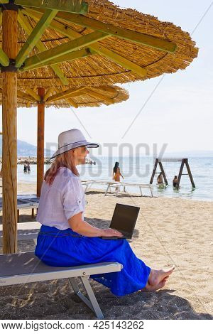 Work From Anywhere. Young Smilyng Woman, Female Freelancer In Straw Hat Working On Laptop On Beach.