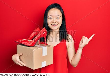 Young chinese girl holding red high heel shoes and package smiling happy pointing with hand and finger to the side 