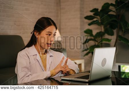 Successful Young Asian Female Doctor With Labcoat And Earphones Doing Video Conference Call For Cons