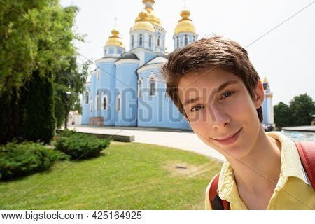 Young handsome 15 years old teen boy looking at camera and happy smiling takes a selfie with smartphone over Mikhailovsky monastery, summer city centr of Kyiv, Europe