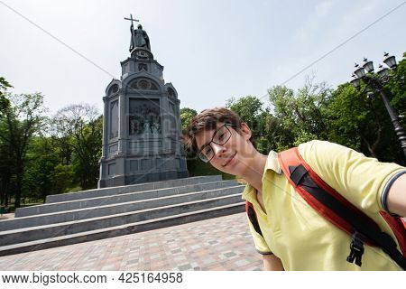 Young handsome 15 years old teen boy looking at camera and happy smiling takes a selfie with smartphone over monument to Vladimir, summer city centr of Kyiv, Europe