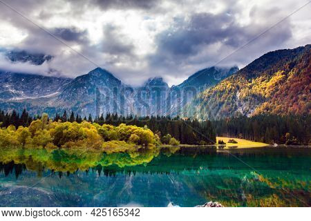 Lake Fuzine in Northern Italy. The Dolomites are covered with clouds. Orange and yellow trees are reflected in the green smooth water. Magnificent colors of autumn. 