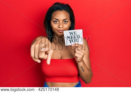 Young african american girl holding we need you paper pointing with finger to the camera and to you, confident gesture looking serious 