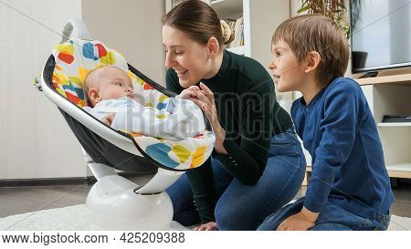 Cute Smiling Baby Boy Rocking In Chair And Looking At Mother Ond Older Brother. Child Development An