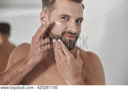 Happy Man With Stylish Beard Applies Antiaging Cream Onto Crowl Feet On Face In Bathroom