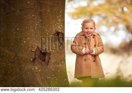 Portrait Of Young Girl Standing By Trunk Of Autumn Tree Playing Hide And Seek In Garden