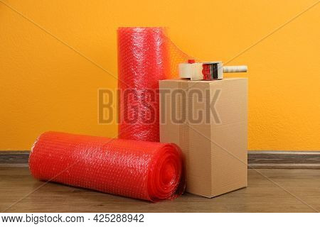 Red Bubble Wrap Rolls, Tape Dispenser And Cardboard Box On Floor Near Orange Wall