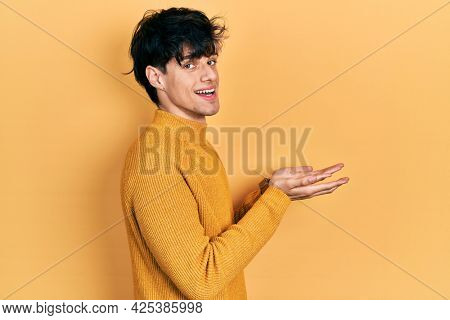 Handsome hipster young man wearing casual yellow sweater pointing aside with hands open palms showing copy space, presenting advertisement smiling excited happy 