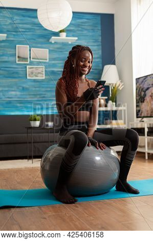 Sporty Athletic Black Woman Relaxing On Stability Ball Checking Social Media Using Smartphone After 