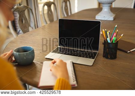 Caucasian woman working in living room at home, writing at table, using laptop, copy space on screen. flexible working from home with technology.
