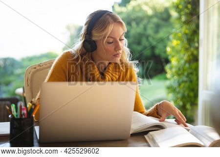 Caucasian woman working in living room at home, wearing headphones and using laptop, reading book. flexible working from home with technology.