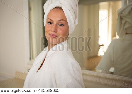 Portrait of smiling caucasian woman in bathroom with towel on head. health, beauty and wellbeing, spending quality time at home.