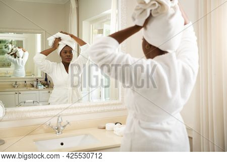 African american woman in bathroom wearing bathrobe, looking in mirror and putting towel on head. health, beauty and wellbeing, spending quality time at home.
