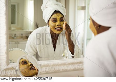 Happy african american woman in bathroom with towel on head, looking in mirror applying face mask. health, beauty and wellbeing, spending quality time at home.