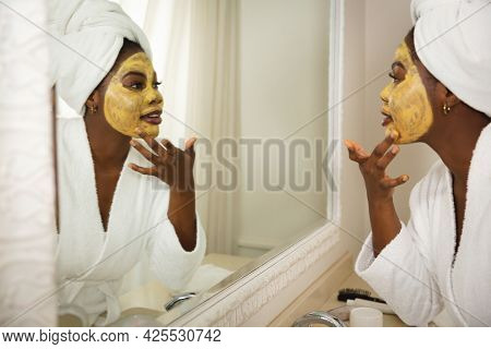 Happy african american woman in bathroom with towel on head, looking in mirror applying face mask. health, beauty and wellbeing, spending quality time at home.