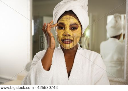 Happy african american woman in bathroom with towel on head, looking in mirror applying face mask. health, beauty and wellbeing, spending quality time at home.