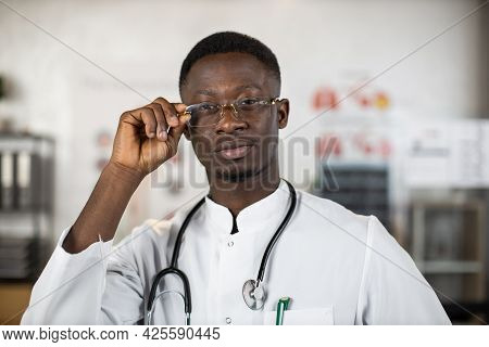 Black Health Care Worker Adjusting His Eyglasses While Posing At Bright Hospital Room. Qulified Doct