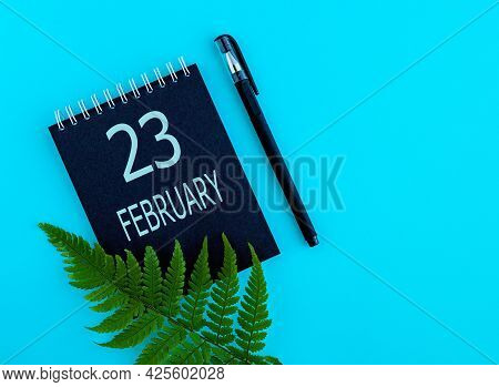 February 23th. Day 23 Of Month, Calendar Date. Black Notepad Sheet, Pen, Fern Twig, On A Blue Backgr
