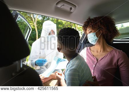 Medical worker wearing ppe suit taking swab tests of african american mother with son sitting in car. medical precautions, lifestyle during covid 19 pandemic.