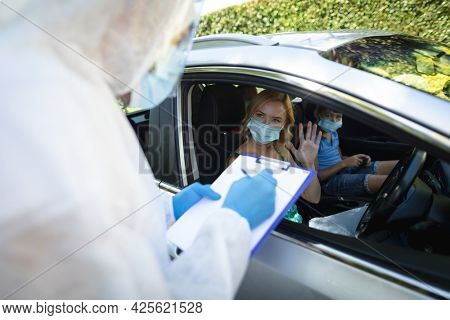Medical worker wearing ppe suit taking notes with caucasian mother with son sitting in car. medical and health precautions, lifestyle during covid 19 pandemic.