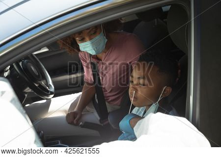 Medical worker wearing ppe suit taking swab tests of african american mother with son sitting in car. medical precautions, lifestyle during covid 19 pandemic.
