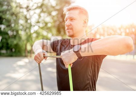 Sportive man guy training with elastic rubber band at the stadium outdoors with sunlight. Athlete male person during workout with additional sport equipmant