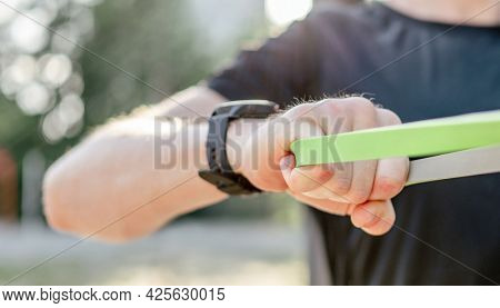 Closeup view of hands of man guy training with elastic rubber band at the stadium outdoors. Adult male person during workout for arms and chest with additional sport equipmant