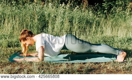 Slim Young Woman Practicing Yoga, Standing In A Plank Pose On A Yoga Mat Outdoors. Girl Yogi In Legg