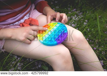 A Little Cute Girl Is Sitting On The Grass With A Pop It - Antistress Toy.