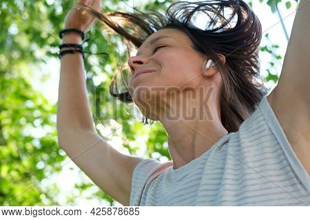 Happy Woman Listens To Music In Wireless Headphones And Dances, With Her Hair Flying Up, On A Summer