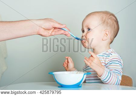 Parent Feeds Child From Spoon Close-up. Handsome Toddler Sitting With Plate Close-up. Baby Complemen