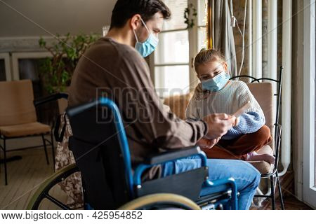 Father With Disability In Wheelchair Using Tablet At Home With Child While Wearing Masks.