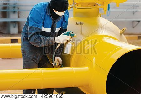 A Painter In Working Clothes Paints A Metal Shut-off Valve For Gasification From A Compressor Gun On
