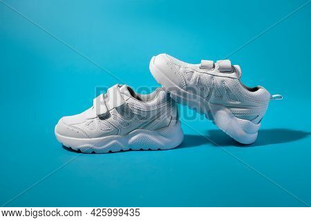 Balancing Two White Childrens Running Shoes On A Blue Paper Background, The Front Of One Sneaker Sta