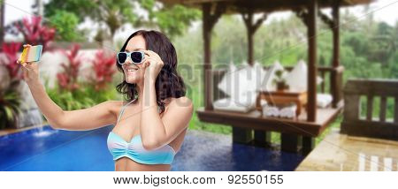 people, technology, summer holidays, travel and tourism concept - happy young woman in bikini swimsuit and sunglasses taking selfie with smatphone over hotel resort background