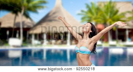 people, summer vacation, travel, tourism and freedom concept - happy woman in bikini swimsuit with raised hands looking up over swimming pool with bungalow and palm trees at hotel resort background