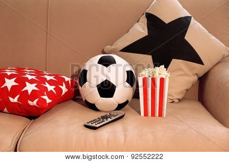 Soccer ball, remote control and box of popcorn on comfortable sofa, indoors