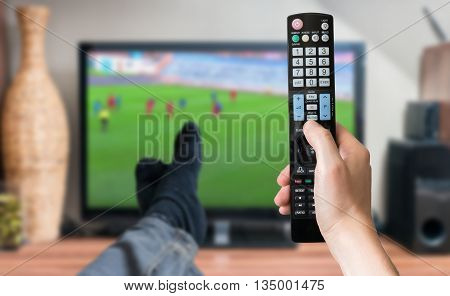 Man is relaxing with legs on table and is watching football match on tv and holds remote controller.