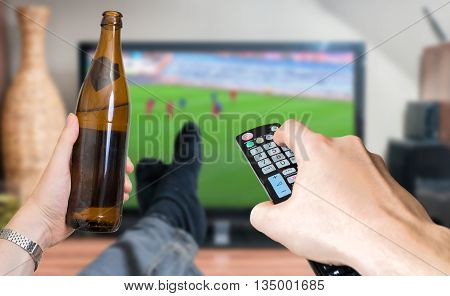 Man is relaxing with legs on table and is watching football match on tv and holds remote controller and beer.