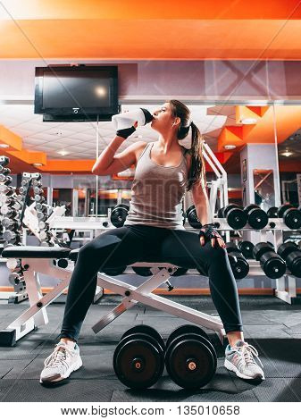 Fitness girl drinking water at gym interior. Break in worout - young sporty woman drink water, sitting near heavy drumbells rack at gym.