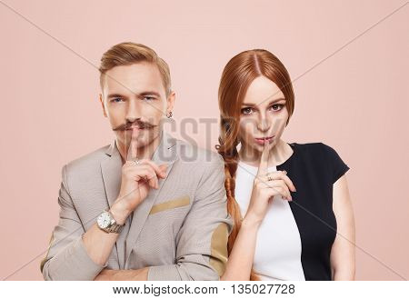 Woman and man keep secret. Couple shows hush sign, adultery, relationship issue, Young beautiful couple isolated at pink