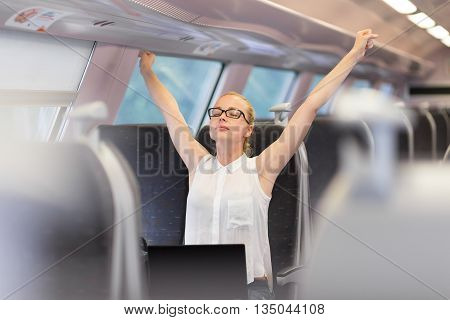 Businesswoman traveling by train, streching her upper body arms reised taking a break from laptop work. Business travel concept.