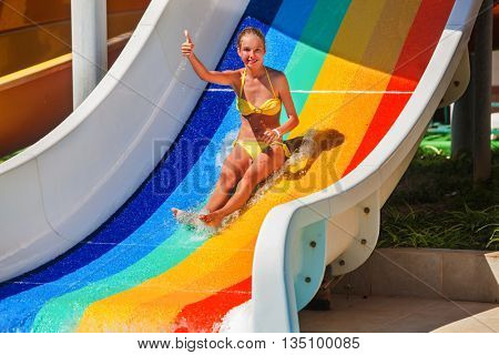 Child teenager wearing bikini slides on water slide and shows thumb up at aquapark . Slide painted in colorful stripes. Summer water park holiday. Outdoor.
