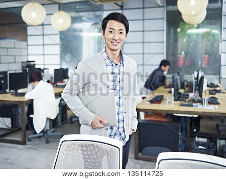 confident young asian entrepreneur standing in own company with laptop computer looking at camera smiling.